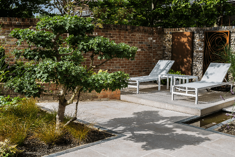 2 shaded sun loungers on a small deck overlooking a paved area and planted border with a small tree