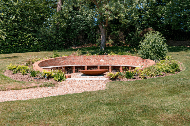 A circular brick seating area with a fire pit in the middle, whcih is surrounded by a planted bed and a lawn.