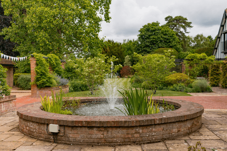 A raised circular brick water feature with a fountain and garden in the background