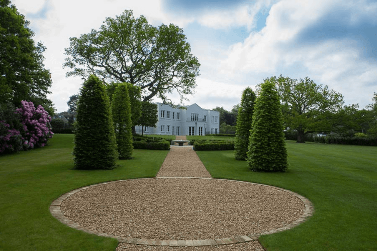A long gravel path cutting through a large lawn with a round section in the fore, leading to a large white house. Either side are some conical topiary trees.