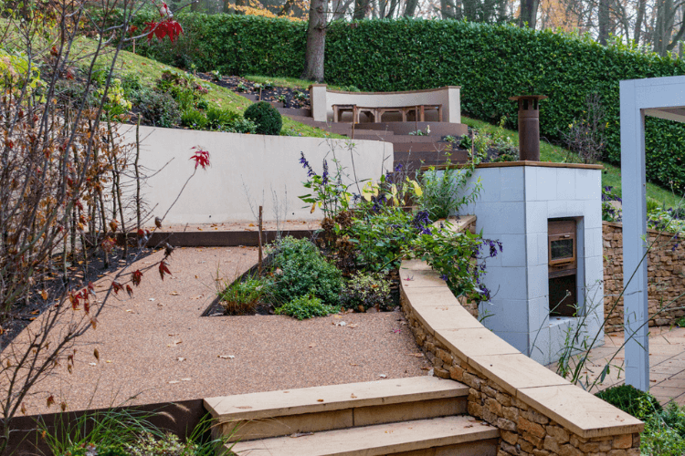 during (4) A white stone outdoor fireplace built into a sandy covered wall which has an inbuilt bed of plants. In the background the garden slopes upwards.