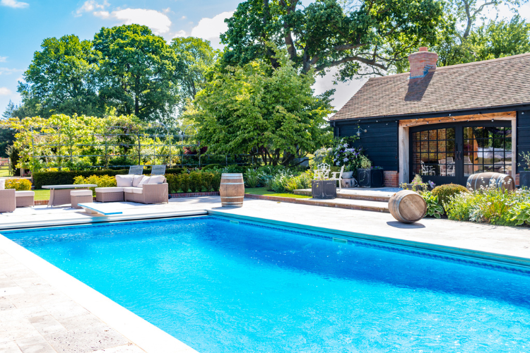 Bright blue rectangular pool with pool house, seating and trees in the background.
