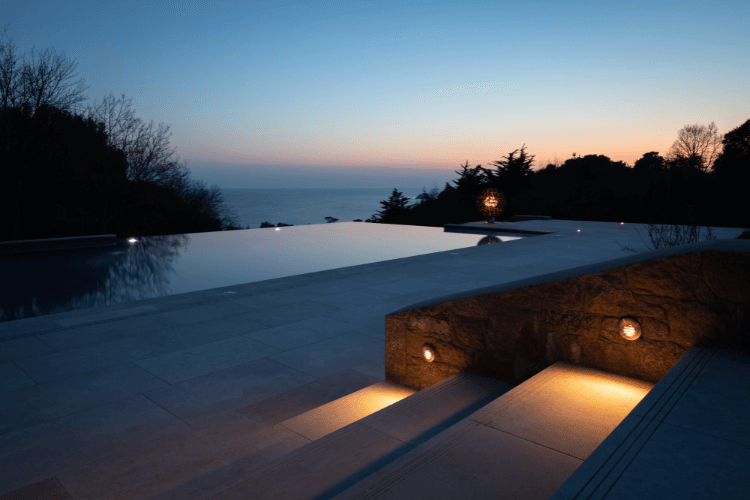 Infinity-edge swimming pool at dusk, carefully integrated into a landscaped setting with subtle lighting and distant coastal views